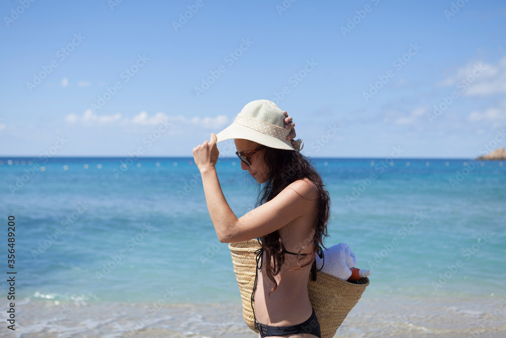 Wanderlust concept of a woman enjoying the summer on the beach in Majorca, the girl has long hair and wears a hat and sunglasses, the woman holds a beach bag or typical Mallorcan basket.