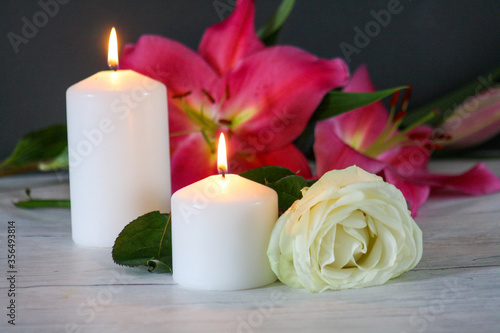 Close up of two pillar burning candles and white rose on dark wooden table with pink lily flowers on the background. Memorial and remembrance concept.
