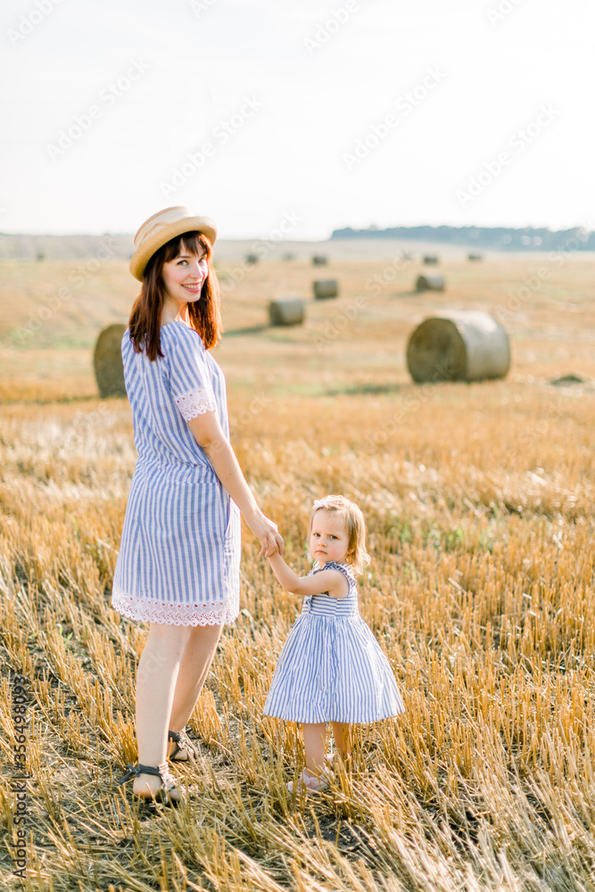 Happy mother and little baby toddler girl holding hands and having fun while walking through summer harvested field with hay bales on the background. Summer sunset. Family walk in the field Happy mother and little baby toddler girl holding hands and having fun while walking through summer harvested field with hay bales on the background. Summer sunset. Family walk in the field