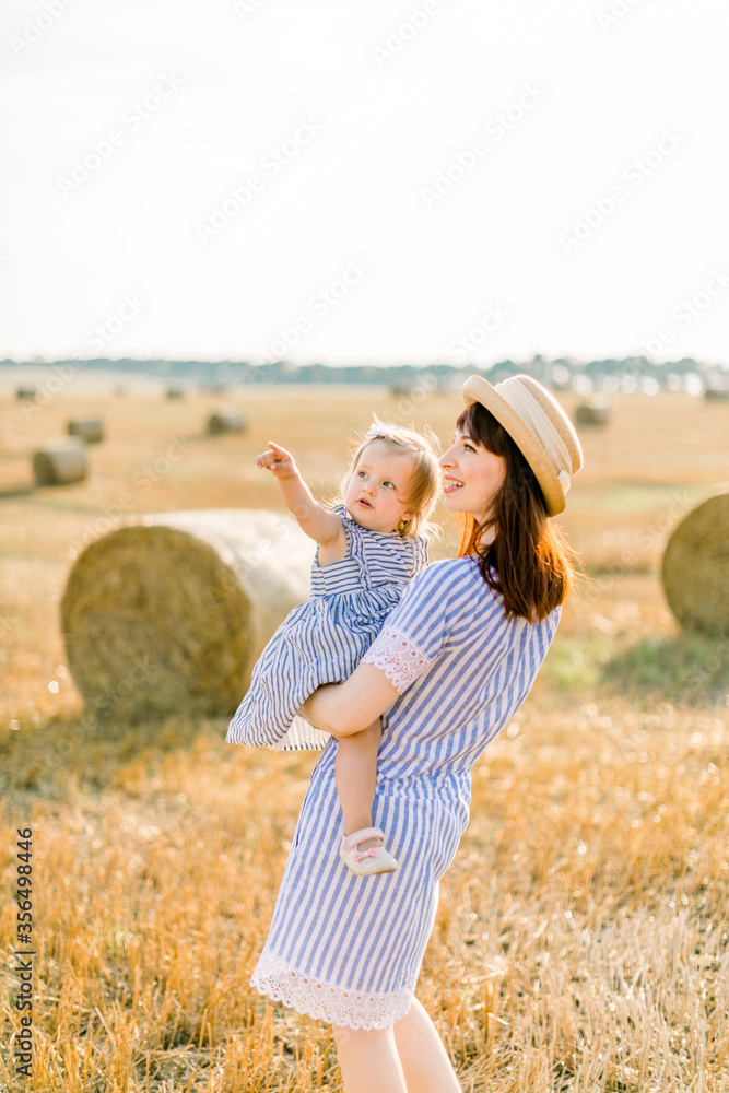 Pretty young mother in straw hat and striped dress holding on hands her cute little baby girl, looking away on the sky, walking in summer field with wheat hay bales on the background Pretty young mother in straw hat and striped dress holding on hands her cute little baby girl, looking away on the sky, walking in summer field with wheat hay bales on the background