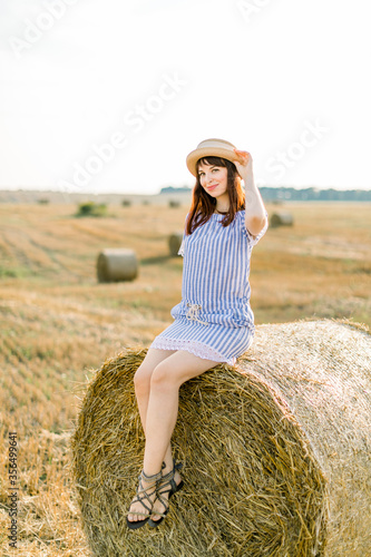 Young brunette Caucasian girl wearing straw hat and blue striped dress, sitting on a hay bale and posing at camera. Summer portrait of young woman in beautiful wheat harvesting field