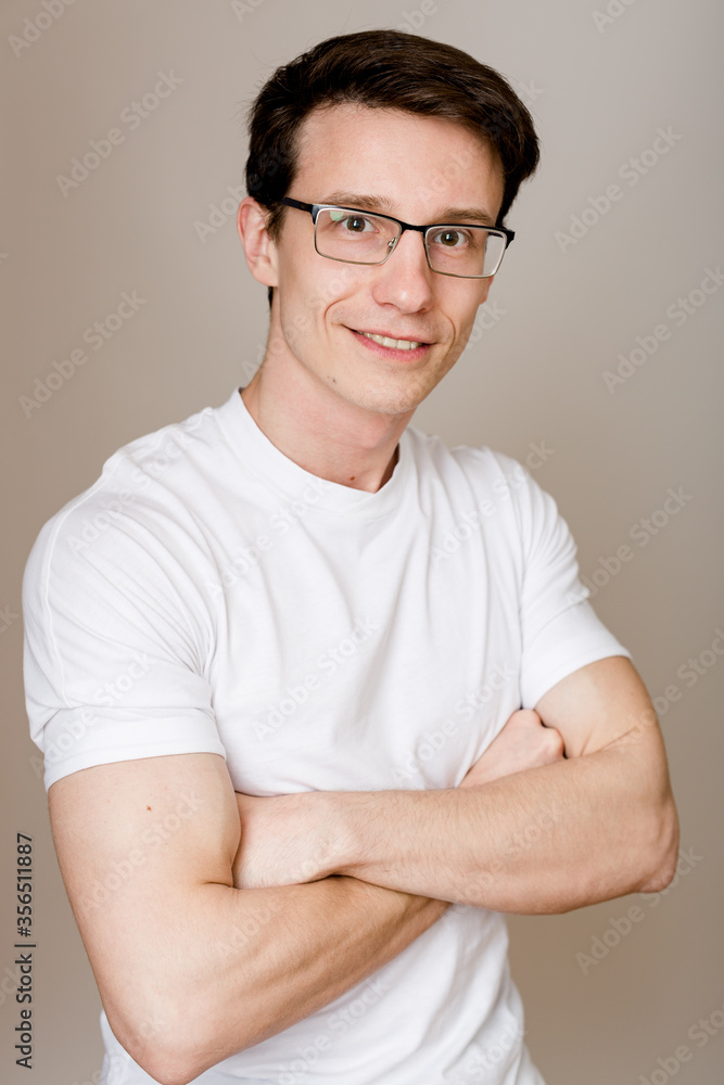 Young confident guy wearing glasses and a T-shirt sits on a grey background with his hands crossed on his chest and a nice, trusting smile looking at the camera. Young confident guy wearing glasses and a T-shirt sits on a grey background with his hands crossed on his chest and a nice, trusting smile looking at the camera.