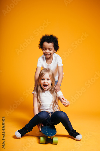 Curly dark-skinned boy rides his little girlfriend on a longboard, and the girl laughs on an orange background