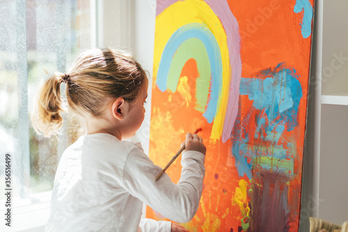 Creative blond girl in casual clothes sitting on window sill against window and painting with paintbrush large multi colored rainbow on orange canvas