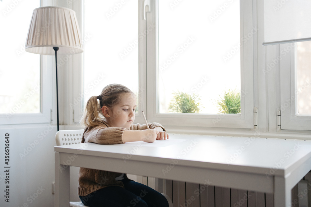 Cute child in casual outfit drawing while sitting at table at light kindergarten Cute child in casual outfit drawing while sitting at table at light kindergarten