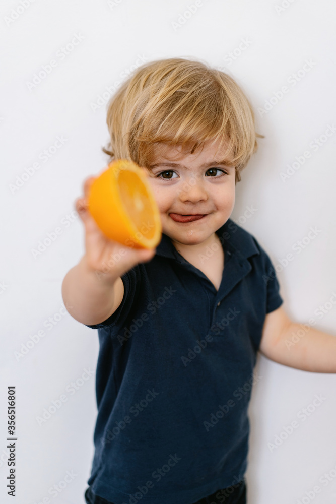 Funny blond little boy in casual clothes stretching offering half of orange towards camera and sticking out tongue with smile while standing against white background Funny blond little boy in casual clothes stretching offering half of orange towards camera and sticking out tongue with smile while standing against white background