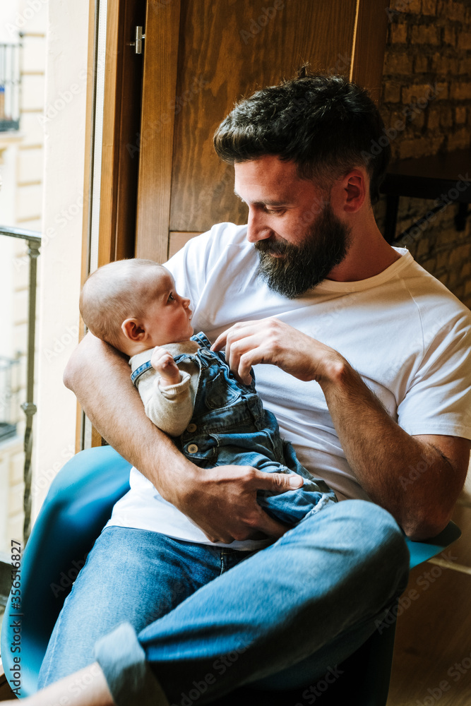 Adult bearded man embracing and communicating with baby while sitting on chair near window at home Adult bearded man embracing and communicating with baby while sitting on chair near window at home