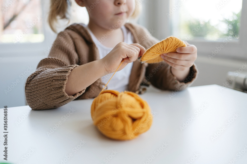 Little girl in casual wear winding woolen threads onto cardboard template to make handmade pompom for hat in cozy living room Little girl in casual wear winding woolen threads onto cardboard template to make handmade pompom for hat in cozy living room