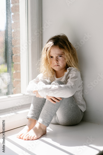 Annoyed little girl with curly hairs wearing gray cozy pajamas looking at the camera with dissatisfaction while sitting on sill with crossed arms