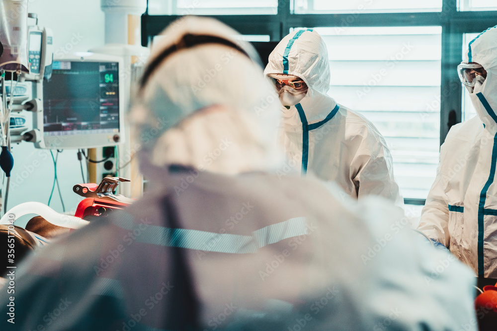Unrecognizable professional doctors in protective uniforms and masks taking care of patient with viral infection while standing in operating room in modern hospital Unrecognizable professional doctors in protective uniforms and masks taking care of patient with viral infection while standing in operating room in modern hospital