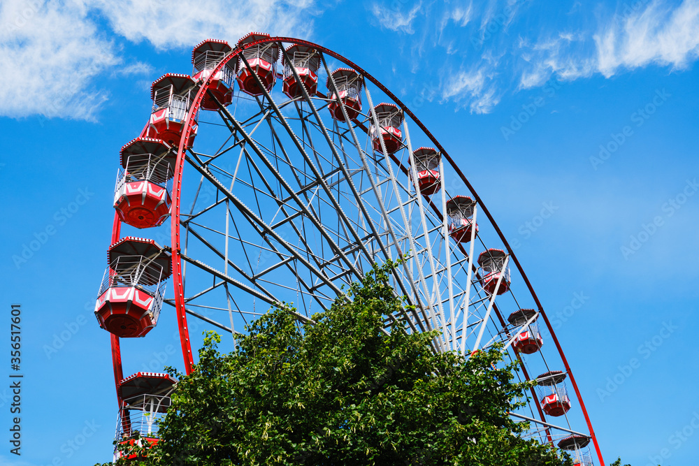 From below of Ferris wheel with red cabins located on amusement park with trees and tower on sunny day with blue sky From below of Ferris wheel with red cabins located on amusement park with trees and tower on sunny day with blue sky