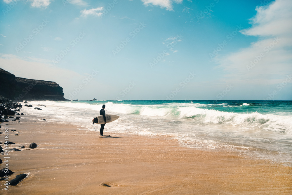 Full body anonymous man with surfboard standing on sandy beach near waving sea water on sunny day on Fuerteventura Island, Spain Full body anonymous man with surfboard standing on sandy beach near waving sea water on sunny day on Fuerteventura Island, Spain