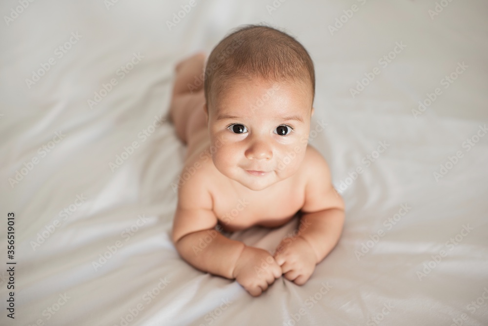 From above adorable infant smiling and looking at camera while lying on soft bed at home From above adorable infant smiling and looking at camera while lying on soft bed at home