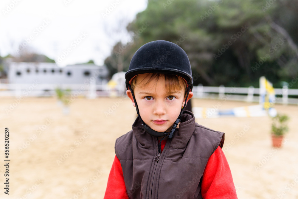 Cute little jockey in helmet and waistcoat looking at camera while standing on blurred background of paddock of equestrian school
