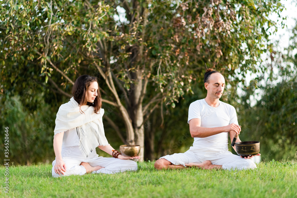 Flexible relaxed couple in white wear sitting in lotus pose holding tibetan singing bowl in hand while meditating and enjoying time together on summer day in park Flexible relaxed couple in white wear sitting in lotus pose holding tibetan singing bowl in hand while meditating and enjoying time together on summer day in park