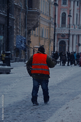 janitor in an orange vest with a shovel on his shoulder walks along a city street during a snowfall