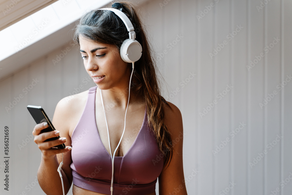 Positive young female in sportswear with headphones browsing smartphone and choosing music for training while practicing yoga at home