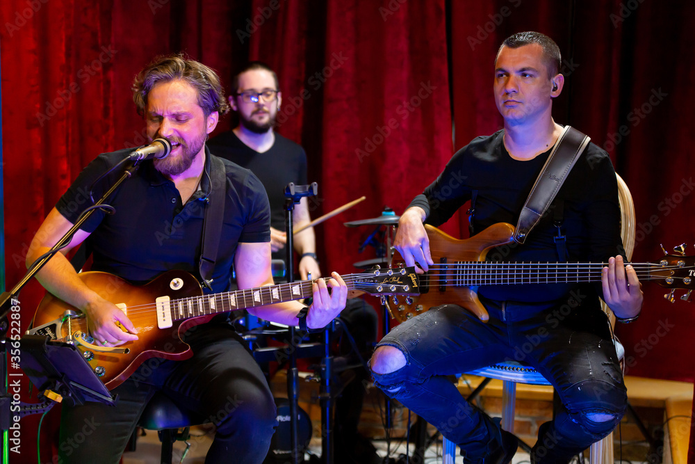 Obraz premium The Band of Musicians playing Electric Guitar, Bass Guitar and Drums during the Concert broadcast Online on Social media platforms in the Bar.