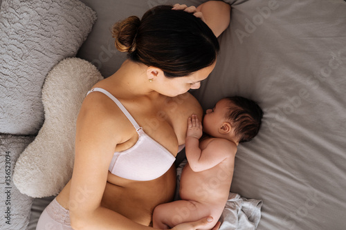 From above of peaceful female in white underwear lying down on comfortable bed and breastfeeding little cute baby