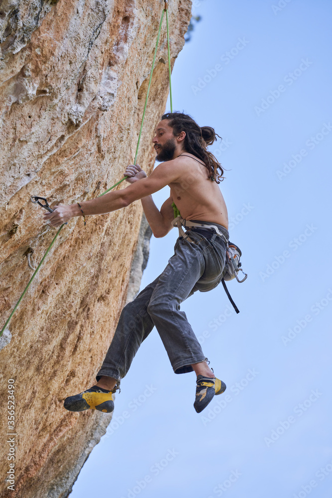 From below side view of shirtless bearded alpinist in safety equipment climbing up on rocky slope of mountain and looking up From below side view of shirtless bearded alpinist in safety equipment climbing up on rocky slope of mountain and looking up