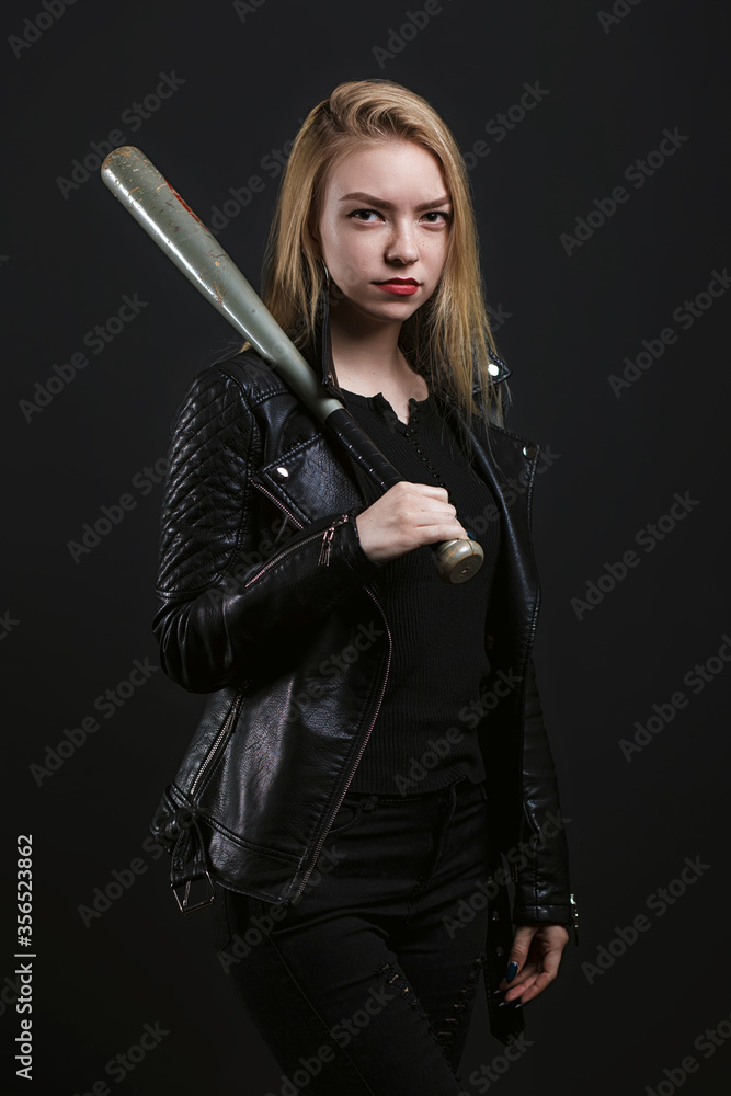 Young beautiful girl in a black jacket is standing with a baseball bat on a black background. Studio photography. Space for text Young beautiful girl in a black jacket is standing with a baseball bat on a black background. Studio photography. Space for text