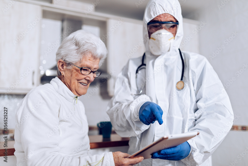 Unrecognizable male medical specialist in protective costume with mask and gloves visiting aged female patient at home during coronavirus lockdown and explaining prescription Unrecognizable male medical specialist in protective costume with mask and gloves visiting aged female patient at home during coronavirus lockdown and explaining prescription