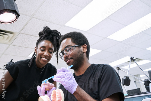 Low angle of happy African American woman with dreadlocks and bearded man smiling and brushing false teeth during work in lab photo