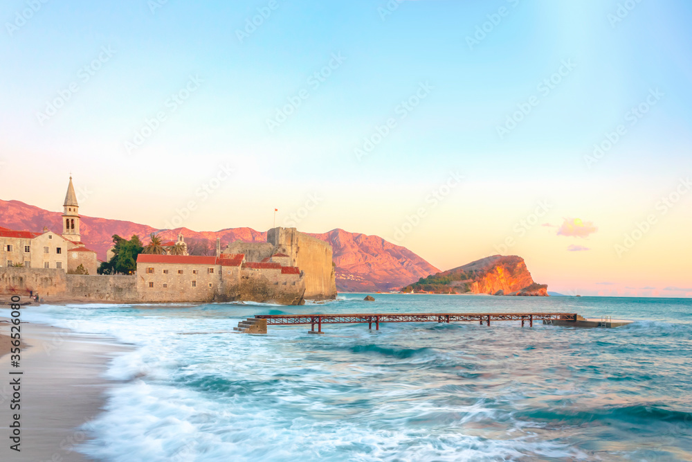 Waves rolling onto the beach and an abandoned pier. Old city in the sunset light. Summer seascape of Montenegro
