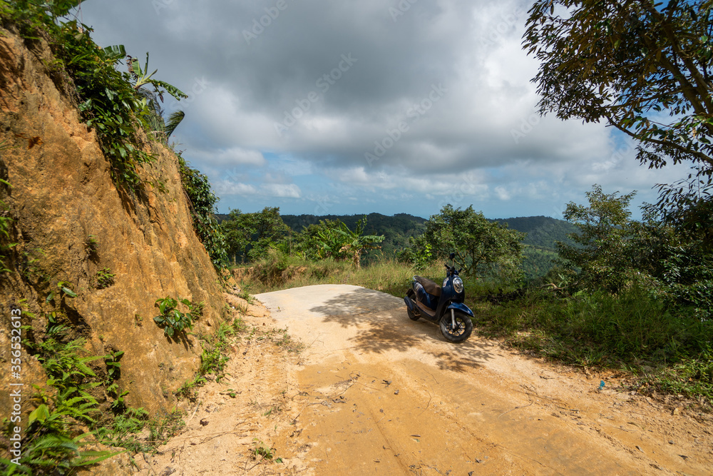 Blue scooter on the dirt road on a hill in tropical jungle with a scenic view on a mountains in a sunny day. Adventure and traveling concept. Blue scooter on the dirt road on a hill in tropical jungle with a scenic view on a mountains in a sunny day. Adventure and traveling concept.