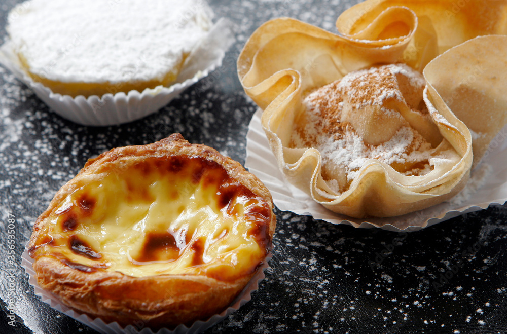 Portuguese egg tart, custard cream pie, named Pastel de Nata. Mix of sweets pastry dessert on dark table with sugar, macro close up view. It's a gourmet typical Portugal recipe. 
