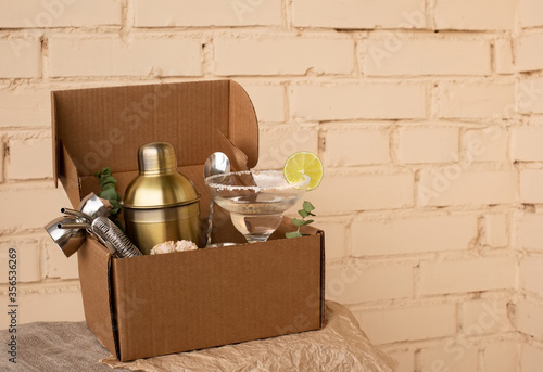 Stay at home bar cocktail party. Classic Margarita cocktail and bartender stuff parcel in a brown craft cardboard box. Gift to mixology lover. Beige brick wall background. Tender retro hipster colors photo