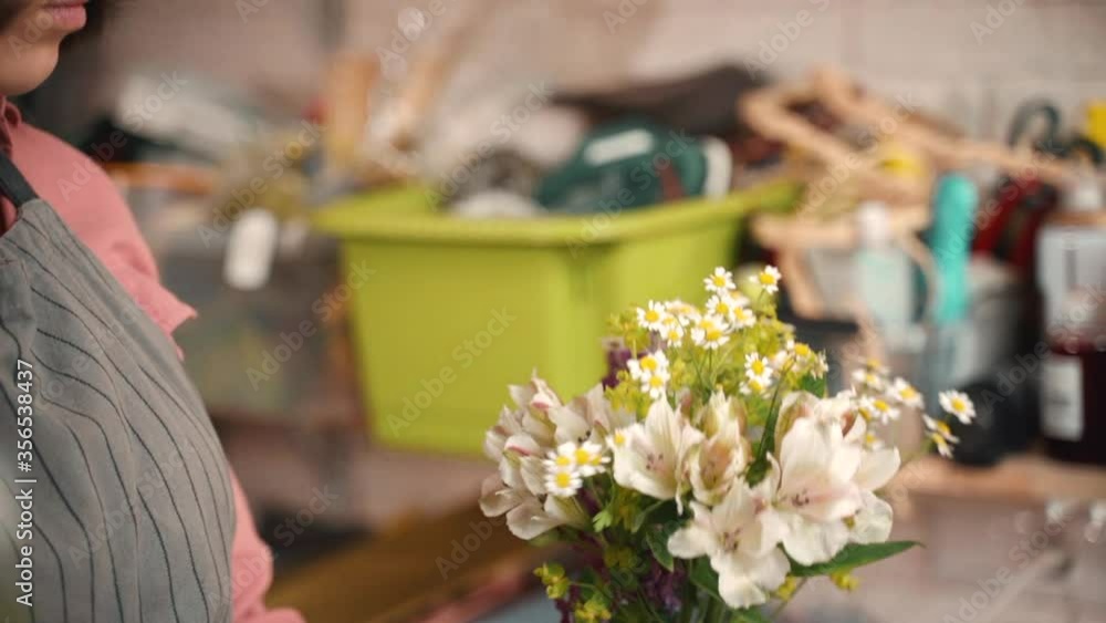 Young woman creating bouquet of fresh flowers while standing in store room. Closeup view of female florist is making romantic composition of blossoms during working day in workshop. Talented person is Young woman creating bouquet of fresh flowers while standing in store room. Closeup view of female florist is making romantic composition of blossoms during working day in workshop. Talented person is