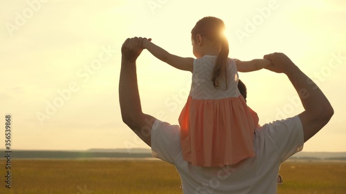 Dad carries on shoulders of his beloved child, in rays of sun. Father walks with his daughter on his shoulders in rays of sunset. child with parents walks at sunset. happy family resting in park.