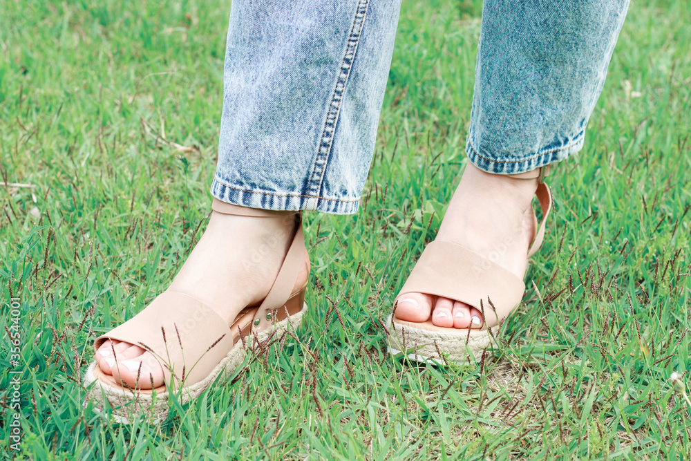 Women's foot in summer brown sandals against background of green grass, concept of summer shoes