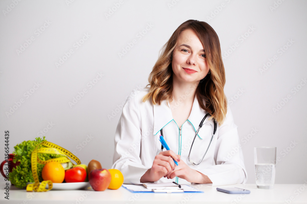 Young dietitian doctor at the consulting room at the table with fresh vegetables and fruits, working on a diet plan