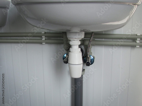 Close-up view of plumbing system under a bathroom sink showing water supply pipes, P-trap drain, and shut-off valves against a white panel wall in a clean, modern utility setting.