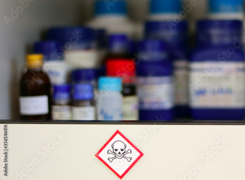 Chemical Hazard Sign pictogram, Globally Harmonized System of Classification and Labelling of Chemicals (GHS) Toxic category with container bottle on shelf cabinet, selective focus blurred background.