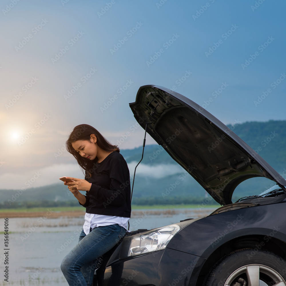 Asian woman with a broken car with open hood.Young woman using mobile phone while looking at broken down car on street