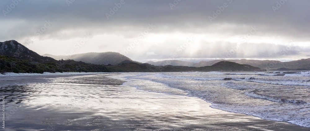 beach ocean water with sand and rocks and green scrub with mountains in the background