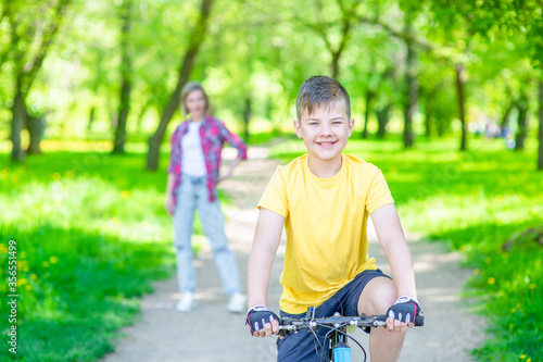 Happy boy rides on a bicycle in summer sunny park. Mom stands behind on the background