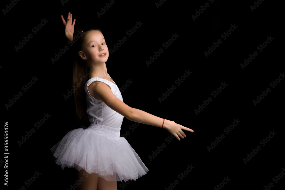 Fototapeta premium Cute little girl in a tutu and pointe shoes is dancing in the studio on a black background.