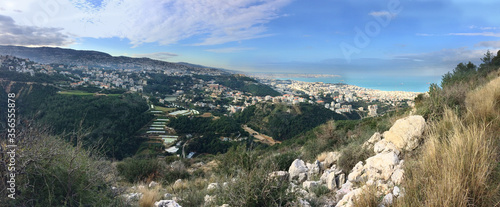 Fototapeta Naklejka Na Ścianę i Meble -  panoramic view of Mediterranean shore and lebanese coast between Kaslik and Beirut, Lebanon, with natural rocks and vegetation in foreground and the village of Aintoura, Lebanon