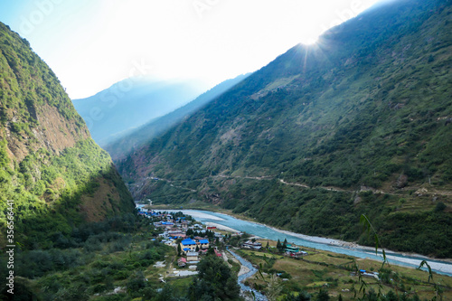 A turquoise river flowing at the bottom of Himalayan valley, surrounding small village Tal, Nepal. There is an Annapurna Circuit Trek going along the valley. Drone capture. High mountain chains