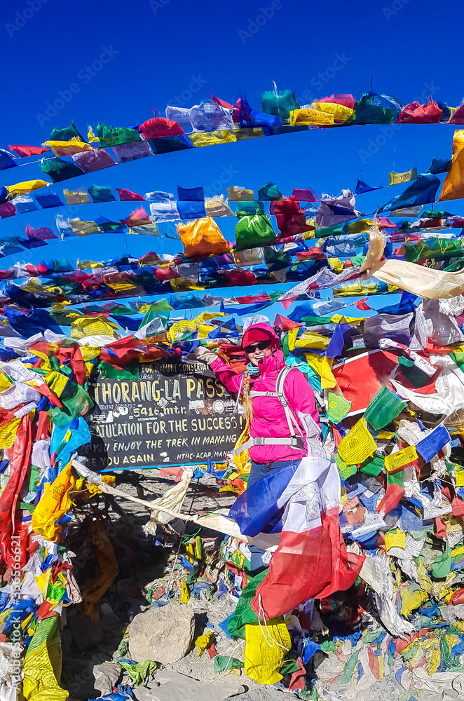 A woman standing between prayer flags at the top of Thorung La Pass, Annapurna Circuit Trek, Nepal. She is very happy. Colorful prayer flags attached to the stone wall, blown by the wind. Clear sky.