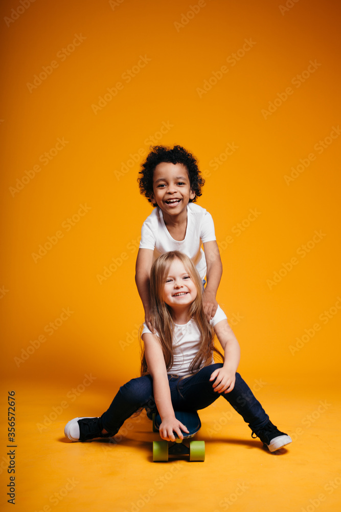 Two young children of different nationalities ride each other on a skateboard and have fun in the studio on an orange background