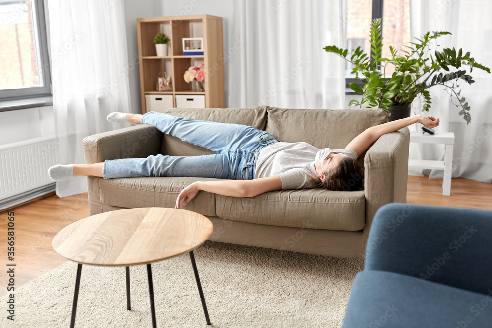 health, safety and pandemic concept - sick young woman wearing protective medical mask lying on sofa at home health, safety and pandemic concept - sick young woman wearing protective medical mask lying on sofa at home