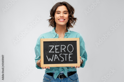 eco living, environment and sustainability concept - portrait of happy smiling young woman in turquoise shirt holding chalkboard with zero waste words grey background photo