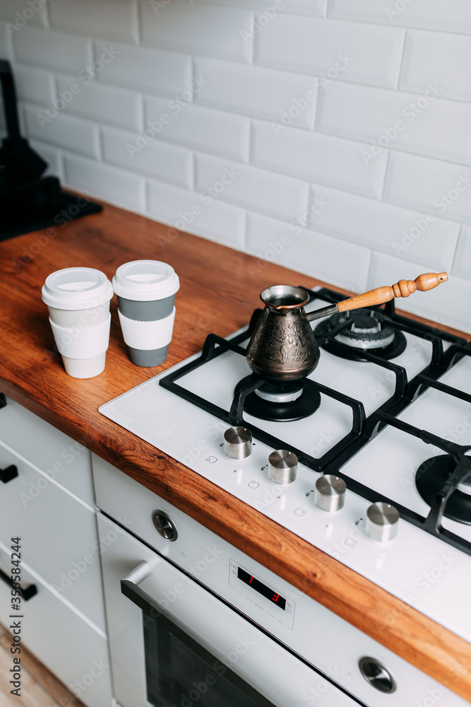 Coffee brewing at home in jezve on gas oven. Coffee cups to go on the wooden table. White kitchen interior. Wooden complete kitchen with gas oven Coffee brewing at home in jezve on gas oven. Coffee cups to go on the wooden table. White kitchen interior. Wooden complete kitchen with gas oven