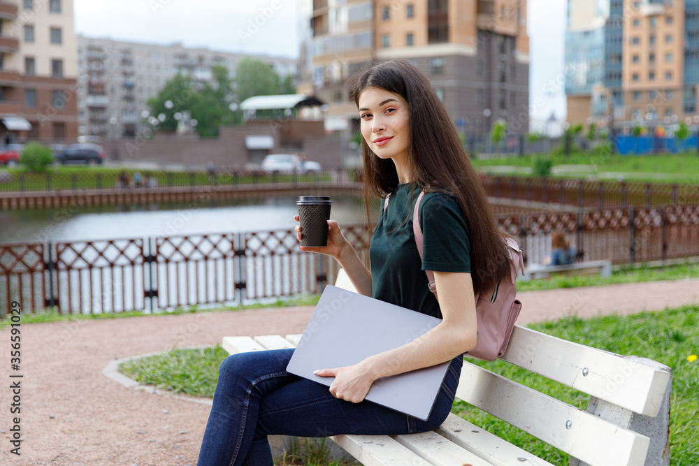 Obraz premium Relaxed young woman in a green T-shirt and with a backpack sitting on a wooden bench, drinking coffee and browsing on her laptop