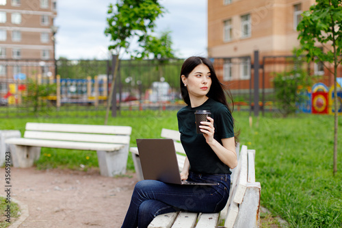 Relaxed young woman in a green T-shirt and with a backpack sitting on a wooden bench, drinking coffee and browsing on her laptop